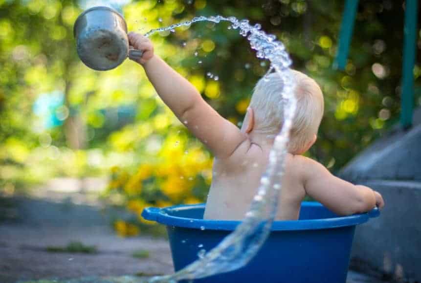 Baby in a blue tub flinging water with a metal cup in a bath where we learn how to clean baby ear wax safely