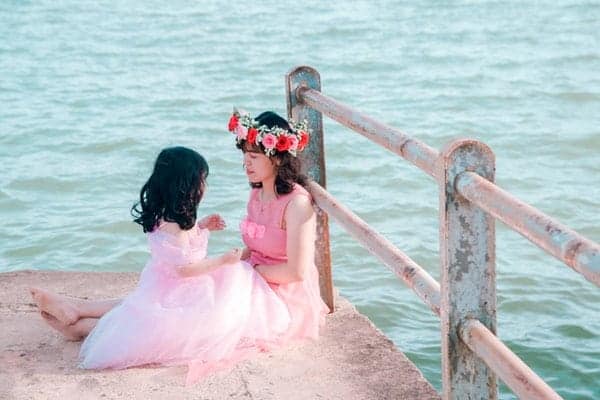 Woman And Girl In Pink Dress Sitting Near Body Of Water