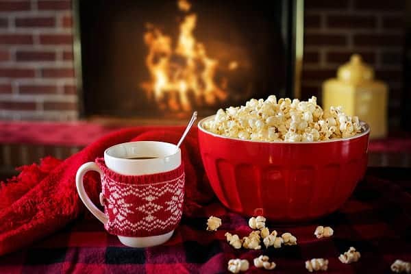bowl of popcorn and a mug of hot chocolate in a knit cozy in front of a fireplace